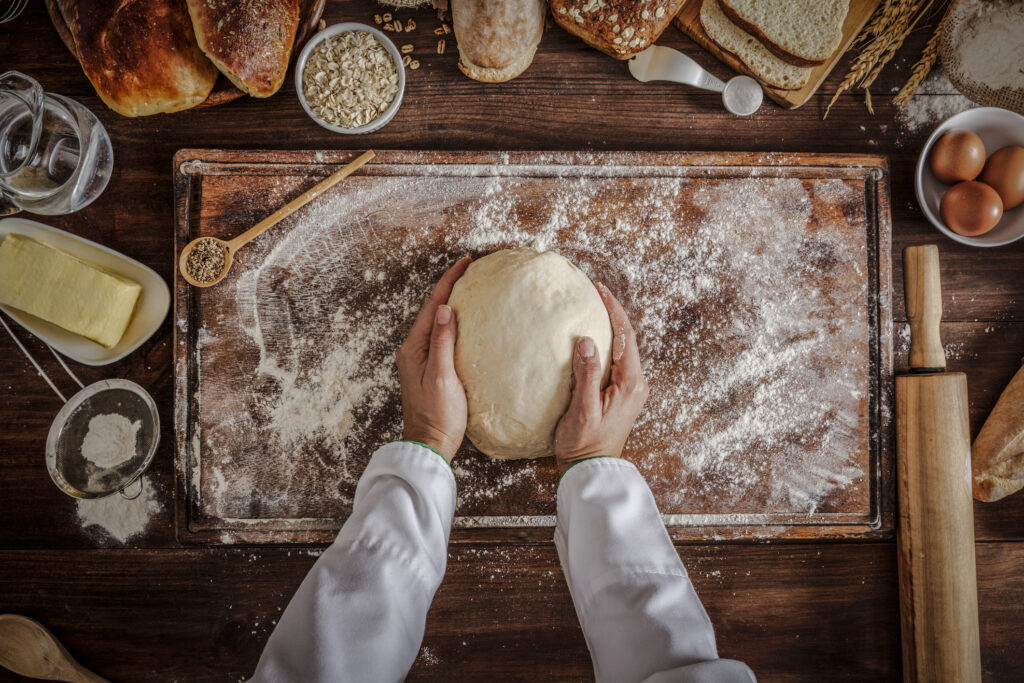 A ball of bread dough being rolled by a chef on a floured board. The dough is surrounded by spoons, seeds and loafs of baked bread.