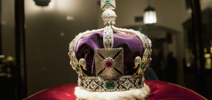 A close-up photograph of a replica of the British Imperial State Crown resting on a red velvet cushion inside a glass display case. The crown features a purple velvet cap, an ermine fur band, and is intricately adorned with diamonds, pearls, and large faux gemstones, including a prominent ruby set in a cross pattée at the front.