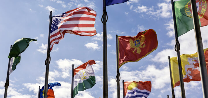 A low-angle view looking up at several international flags waving from tall metal flagpoles against a sunny blue sky with light, patchy clouds. Prominently featured on the upper left is the United States flag. To the right, the flags of Montenegro (red with a gold border and coat of arms) and Portugal (green and red with an armillary sphere) are visible. Below them fly the flags of Tajikistan, Kyrgyzstan, and Bhutan, among others, creating a colourful display of global representation.