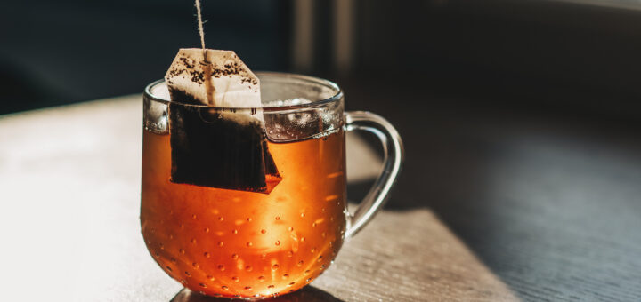 Close-up of a clear glass mug filled with steeping amber-colored tea sitting on a wooden surface. A tea bag hangs from its string into the hot water, with visible dark tea clouds swirling into the liquid. Bright natural sunlight streams in from the side, illuminating the tea and casting a warm shadow against a dark, out-of-focus background.